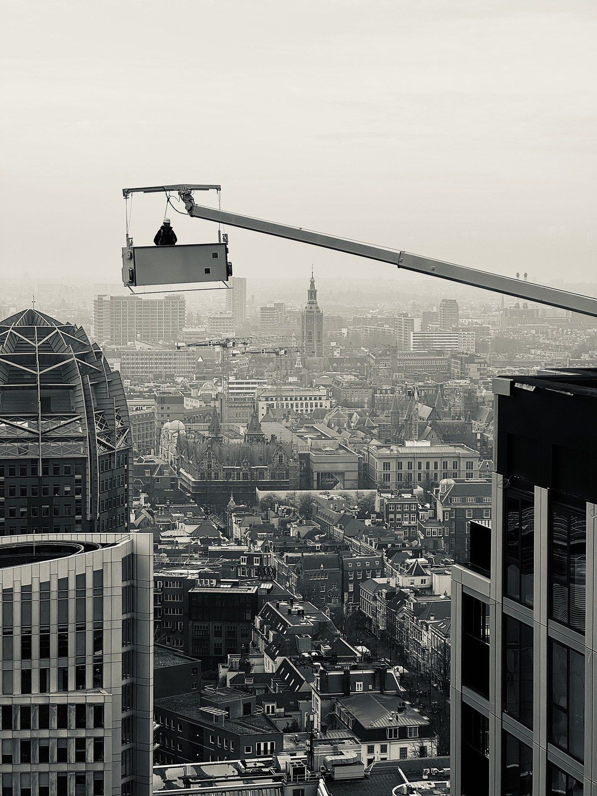 A maintenance cradle hanging from a roof arm above a city, in black and white, a Wikimedia Commons photograph in Michael Stern's encyclopedia