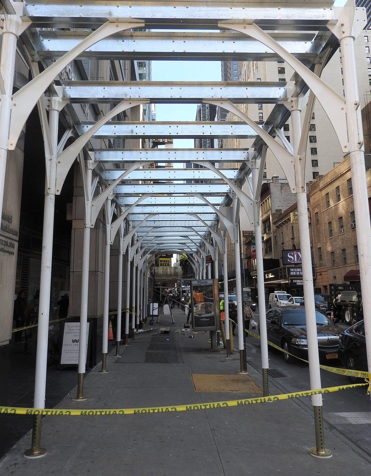 The steel frame of a sidewalk shed over a city pavement before its deck is laid, a Wikimedia Commons photograph in Michael Stern's encyclopedia